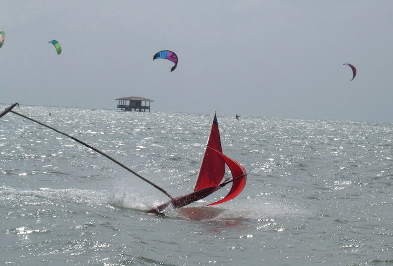A red-sailed jong capsizing and throwing spray, kitesurfers and a stilt hut visible on the horizon behind it