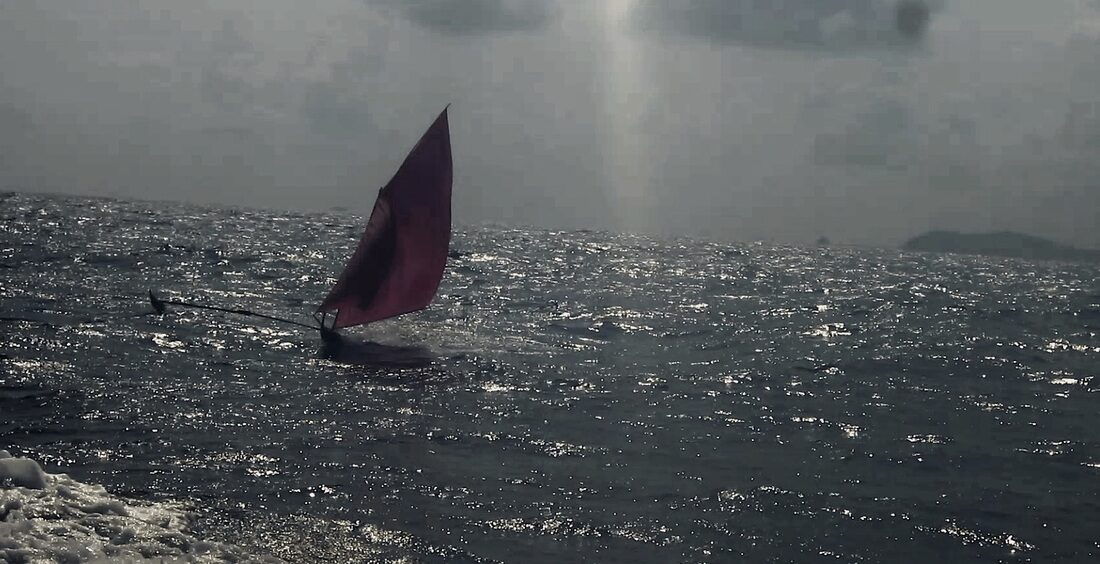 A jong with a dark red sail catches the last light on open water, glare from a low sun on the sea