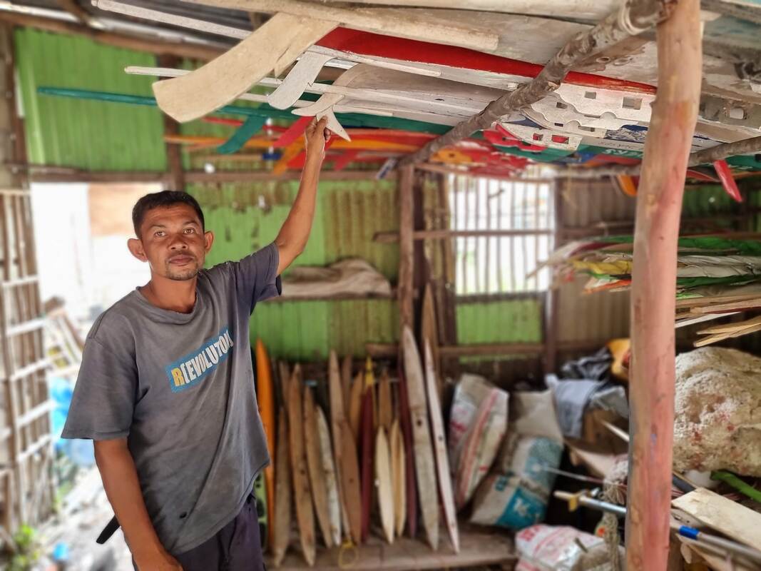 A man in a workshop pointing up at rows of painted outrigger floats and hull pieces stored under a tin roof