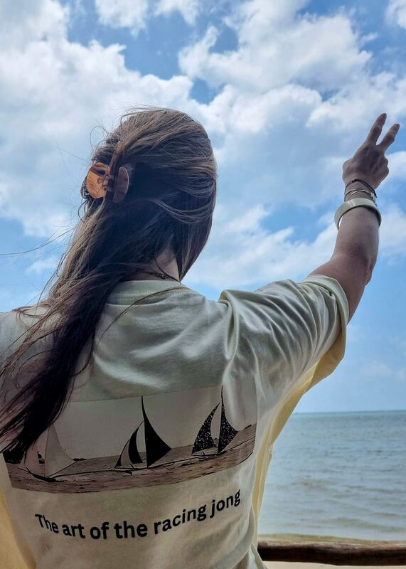 A woman raising a peace sign to the sea, wearing a Jong School t-shirt printed with racing jongs and the slogan 'The art of the racing jong'