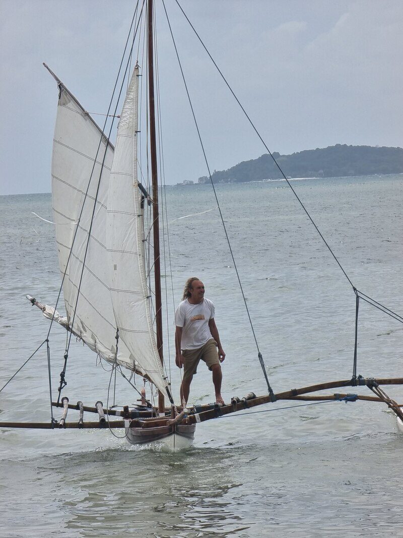 A man in a white shirt standing on a sampan layar's outrigger boom, white sails raised, an island visible on the horizon