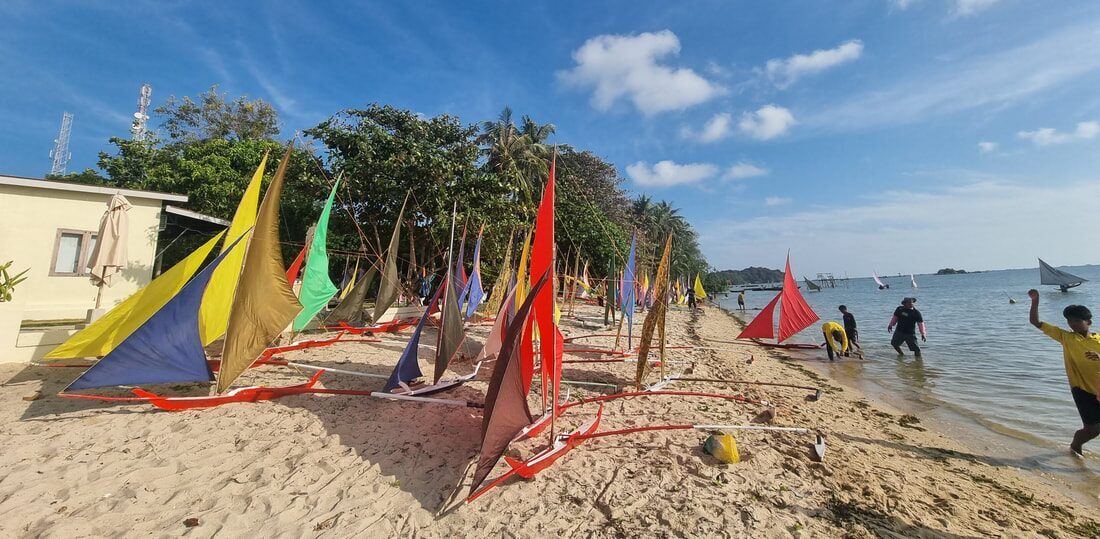 A lineup of colorful jongs on the beach in daylight, people gathered around them