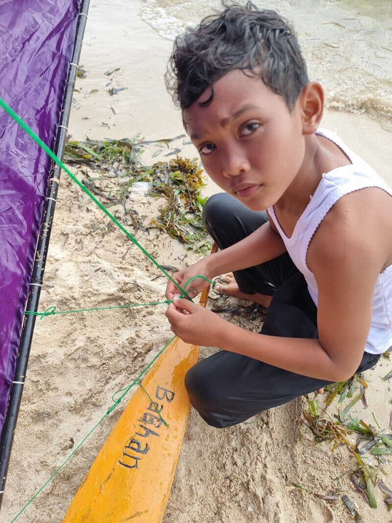 A boy kneeling to tie rigging on a yellow hull named 'Belhan,' waves lapping the beach behind him