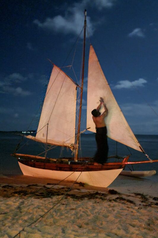 A man adjusting the sail rigging of a sampan layar beached at dusk, its white sails lit against the darkening sky