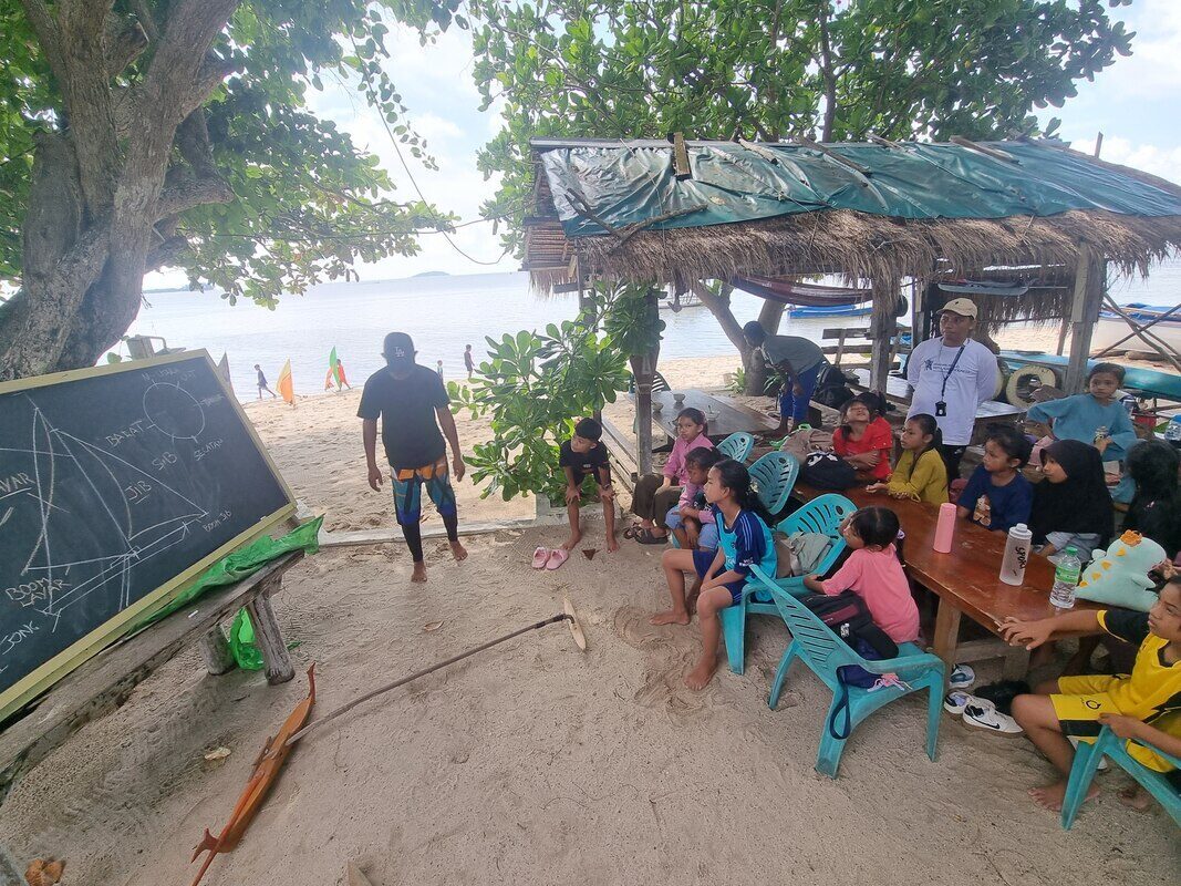 A teacher lecturing at a chalkboard diagram of a jong's parts, kids listening at a table under a thatched shelter by the beach