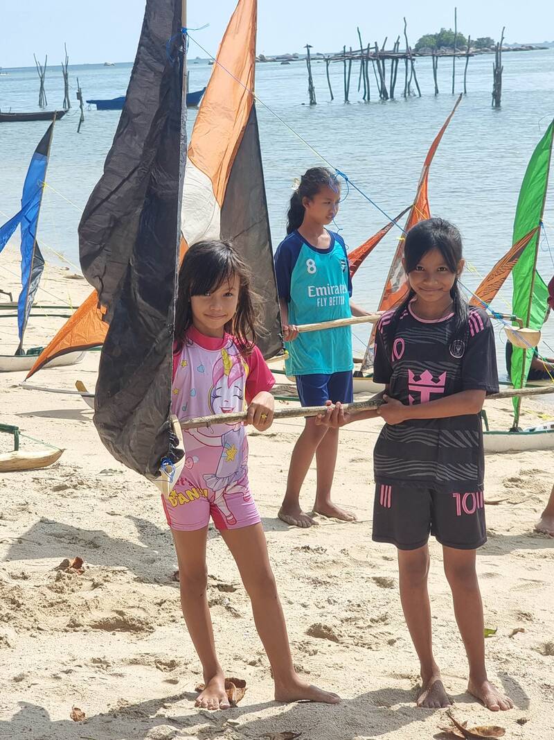 Three girls holding a jong's sail spar on the beach, orange and black sails and old fishing posts in the water behind them