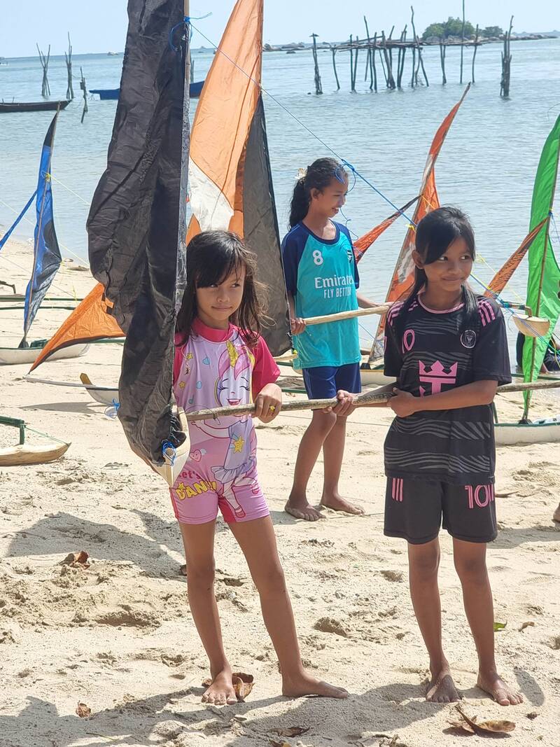 Three kids holding jong sail spars on the beach, boats and old fishing posts in the water behind them