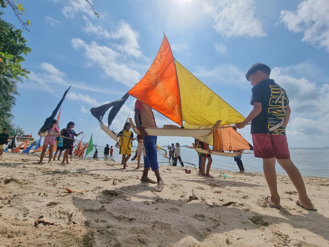 A boy carrying a jong with orange and yellow sails down the beach among other kids and boats