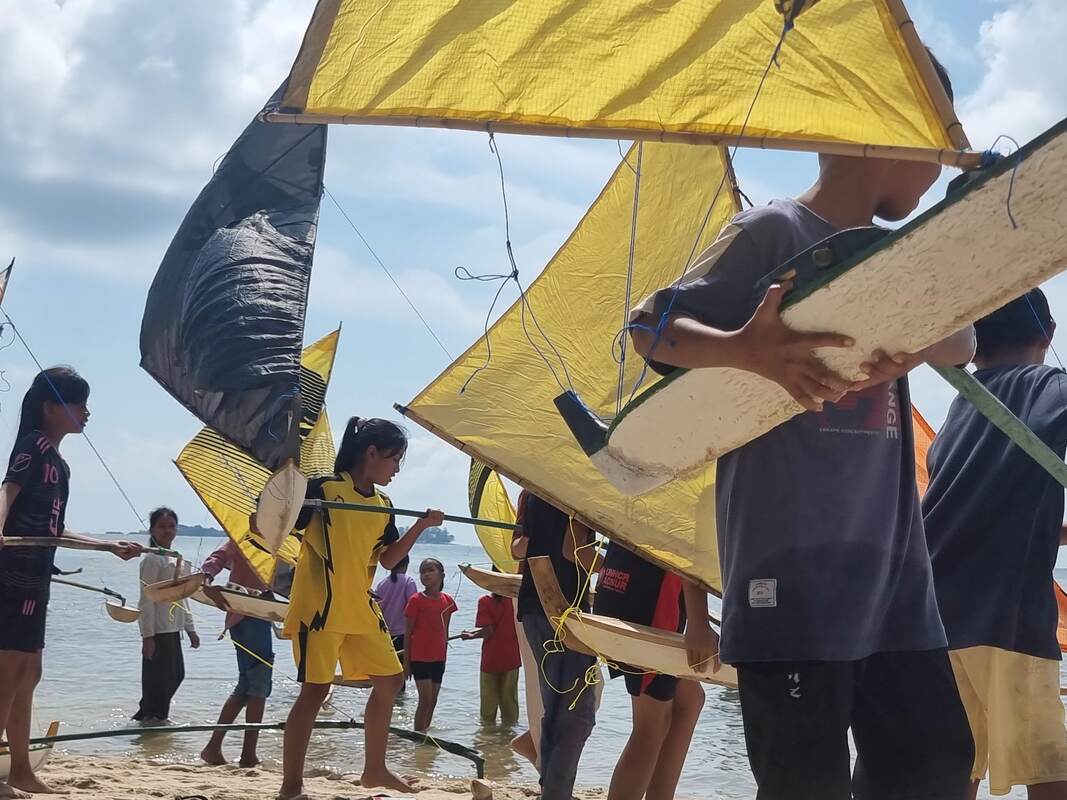 Kids carrying jongs with yellow and black sails through the crowd toward the water