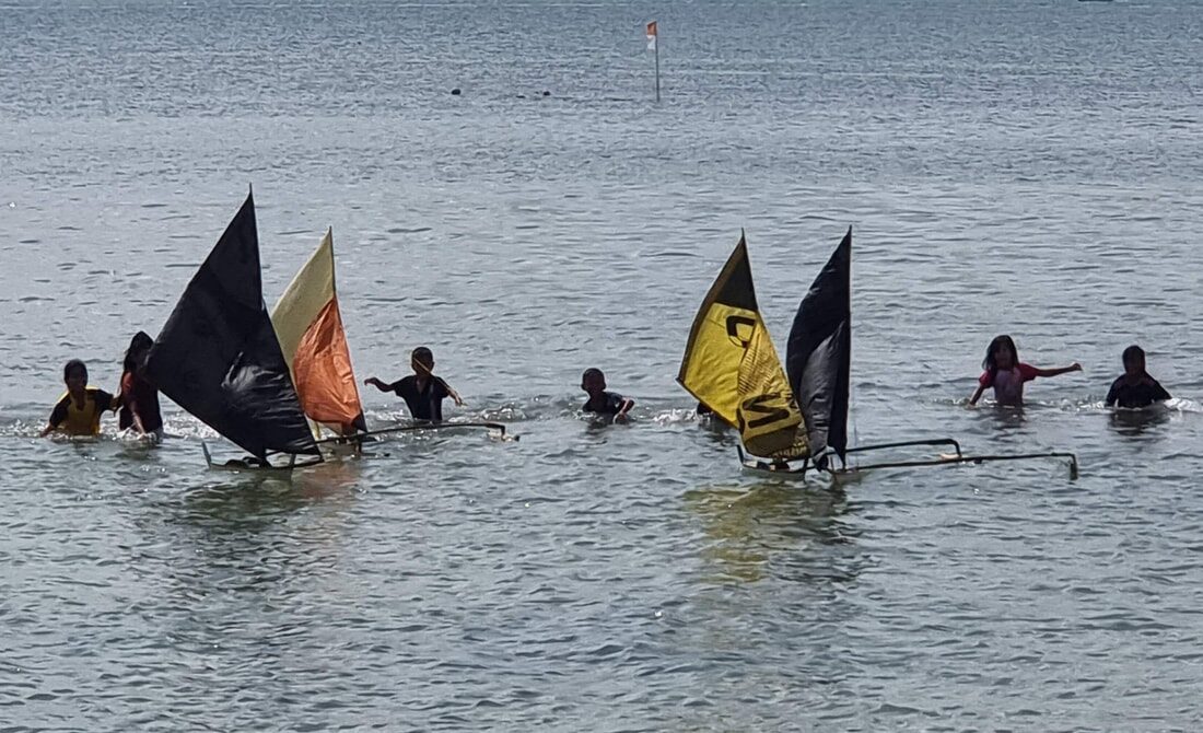 Kids wading chest-deep with black and yellow-sailed jongs, a race marker flag in the distance