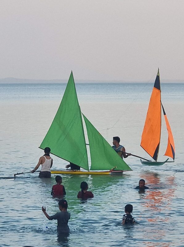 A green and orange jong sailing close to shore, kids in the water nearby