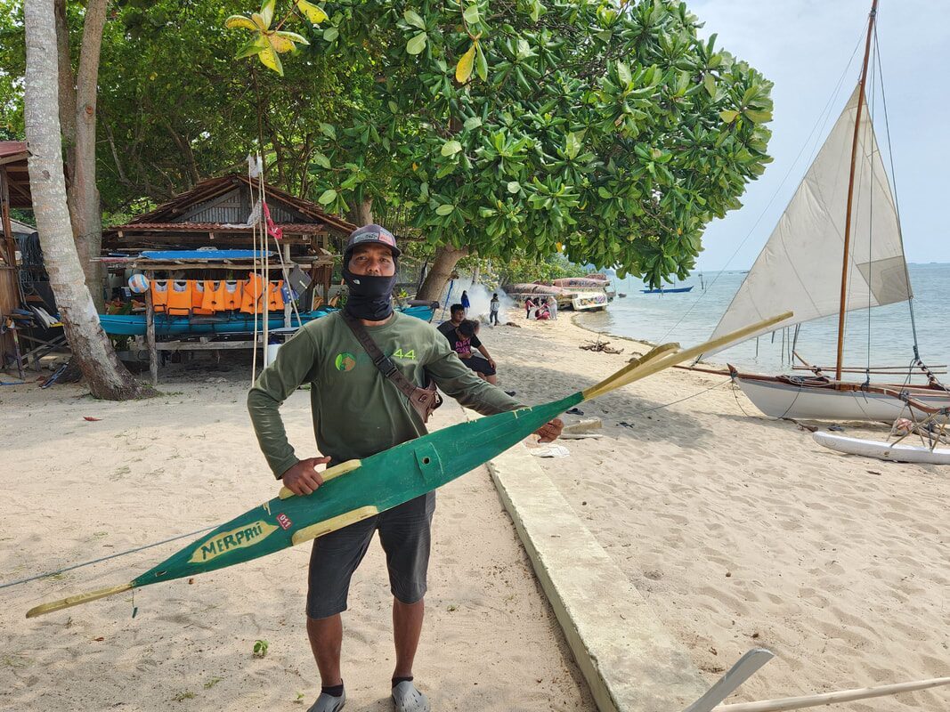 A player holding a green outrigger float named 'Merpaii,' a white-sailed jong resting on the sand behind him