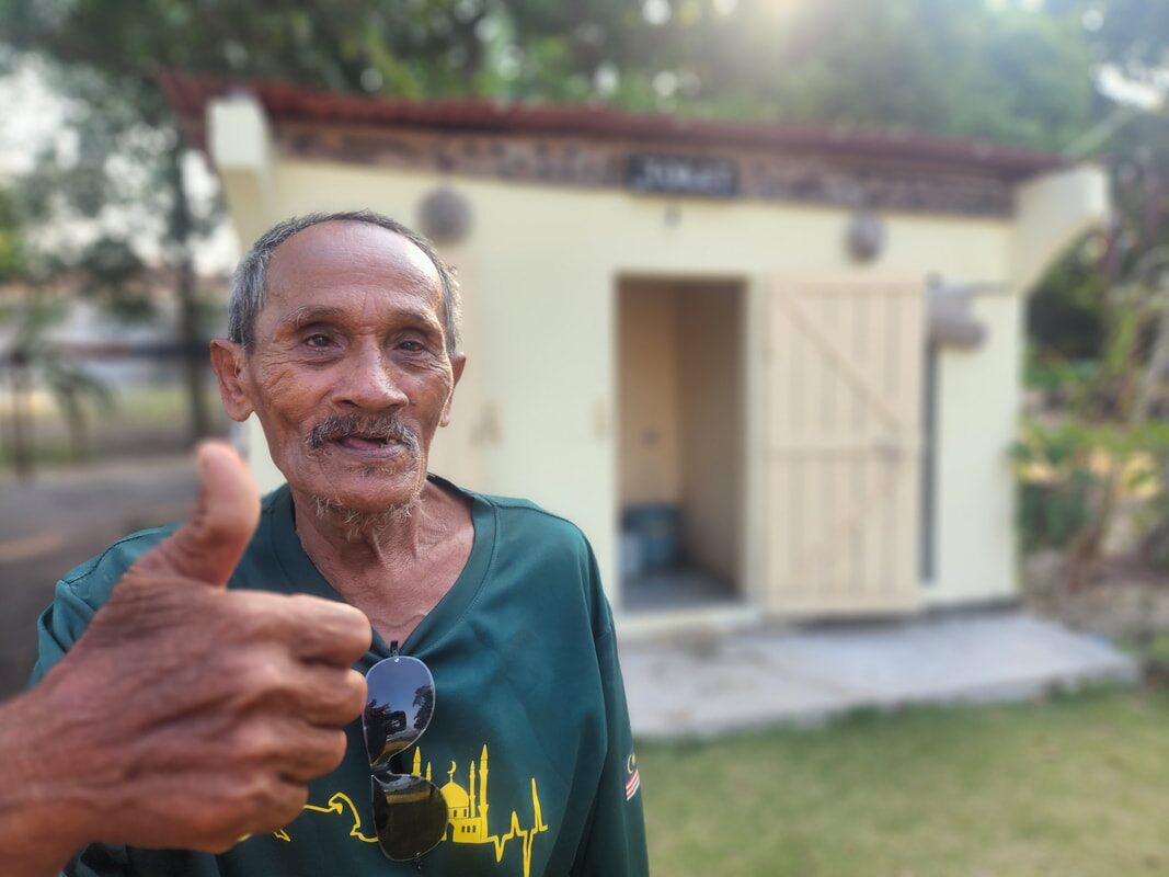 Portrait of an older man giving a thumbs up outside a small building
