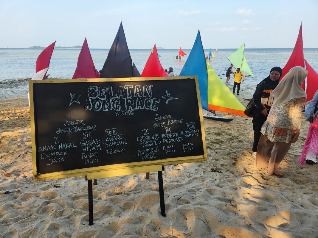 A chalkboard results sign for the Selatan Jong Race 2026 listing category winners, with racing jongs and beachgoers behind it