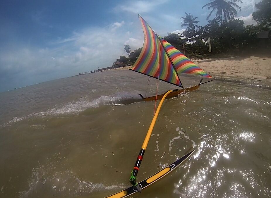 A rainbow-sailed jong riding waves toward the beach at Teluk Bakau, palm trees along the shore