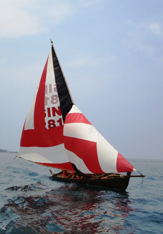 A traditional sampan layar under a red-and-white striped sail, crew visible aboard, sailing open water