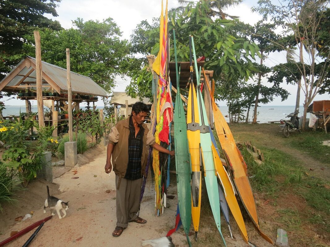 A man standing beside a rack of painted outrigger floats and jong hulls in a garden workshop, a cat nearby