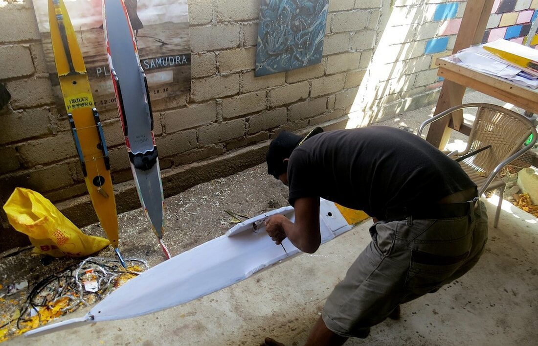 A man in a cap bent over sanding a jong hull outdoors, with painted outrigger floats and posters propped against a brick wall behind him