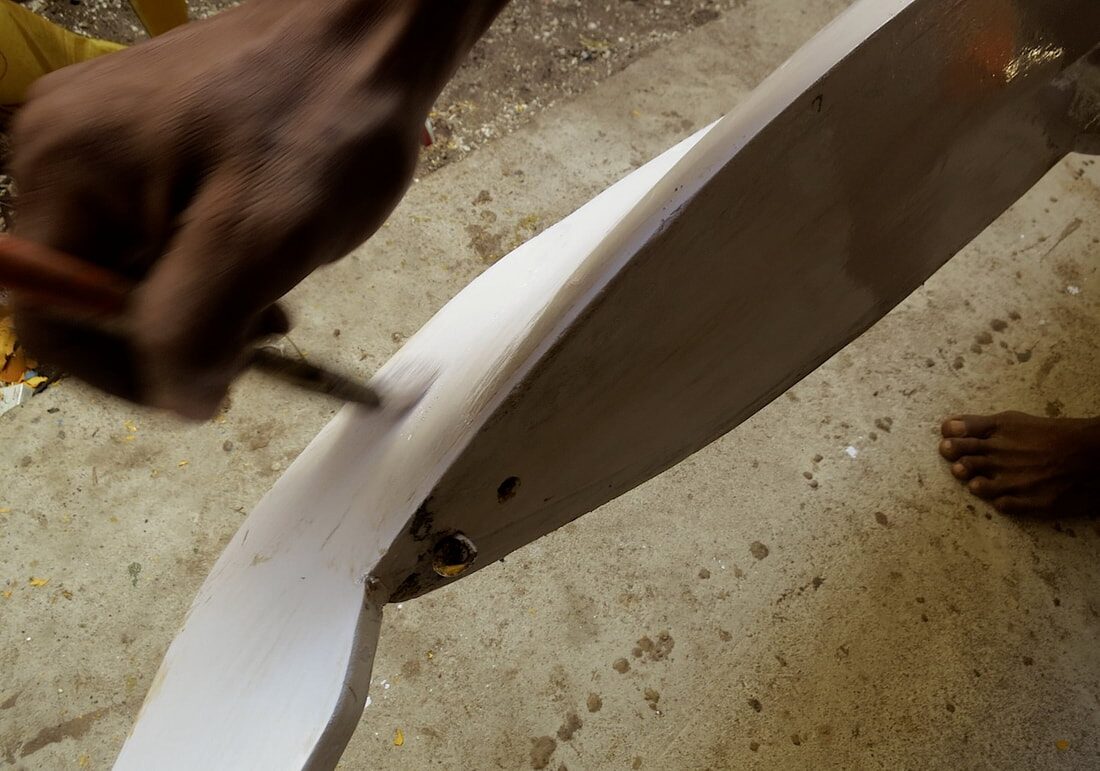 Close-up of hands sanding a white-painted jong outrigger float, a bare foot resting nearby