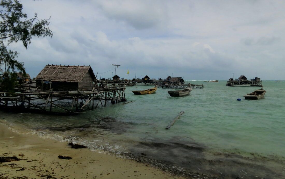 A stilt-house sea village seen from the beach, small wooden boats anchored in the shallows