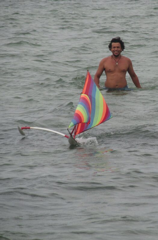 A jong with a rainbow-striped sail in the water, a man wading behind it