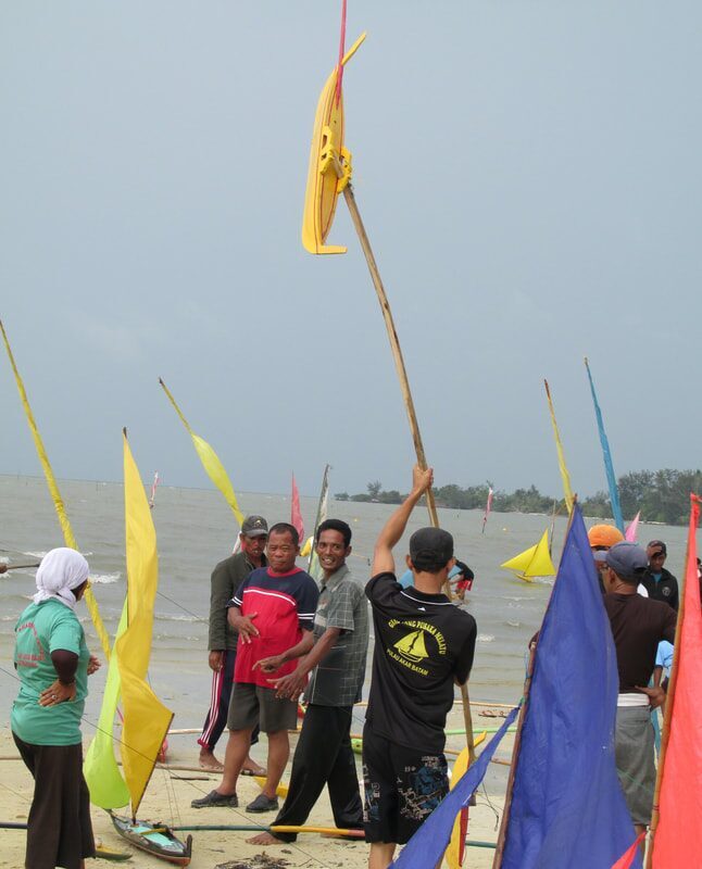 A jong's tall wooden mast and sail being hoisted on the beach at Dompak, racers gathered around