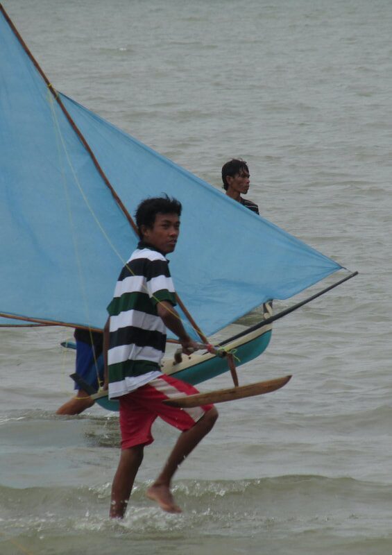 Two men wading a jong with a pale blue sail out into the water at Dompak