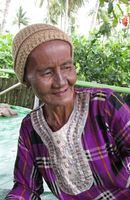 Portrait of an elderly woman in a woven cap and purple batik top, smiling at a race-day gathering