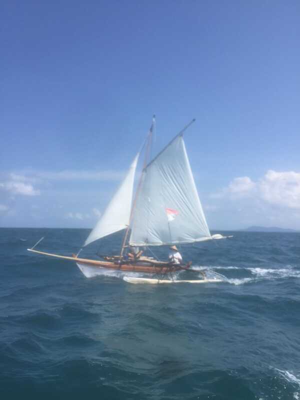 A traditional outrigger sailboat with white sails and a small red-and-white flag, heeling as it sails choppy blue water
