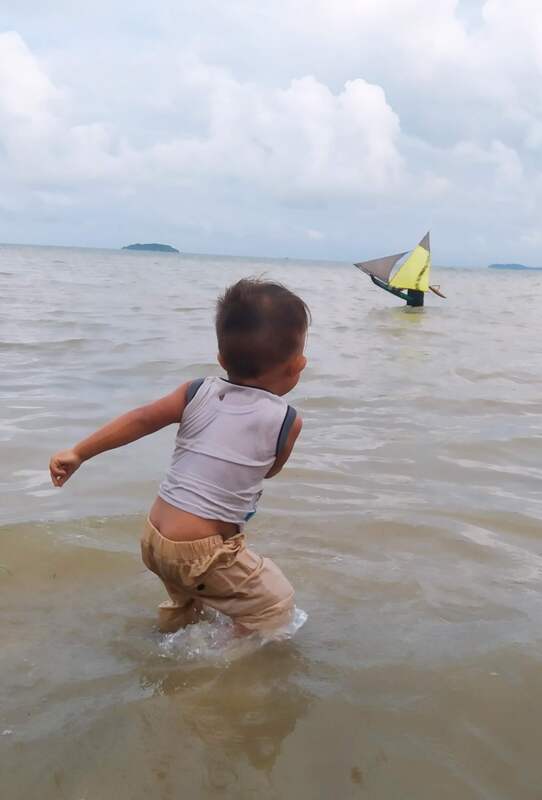 A toddler wading out toward a small jong with a yellow and grey sail