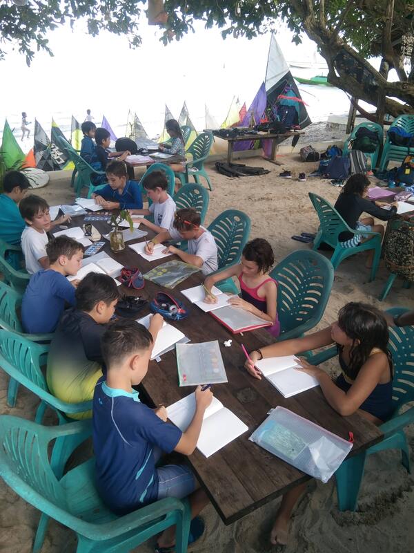 Kids seated at a long table under the trees, writing in workbooks during a Jong School lesson, sails drying nearby