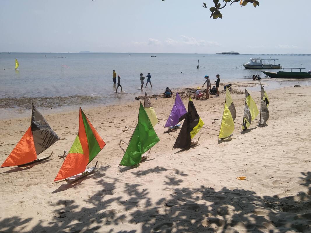 A line of jongs in orange, green, purple and yellow sails staked along the beach, kids playing in the shallows behind them