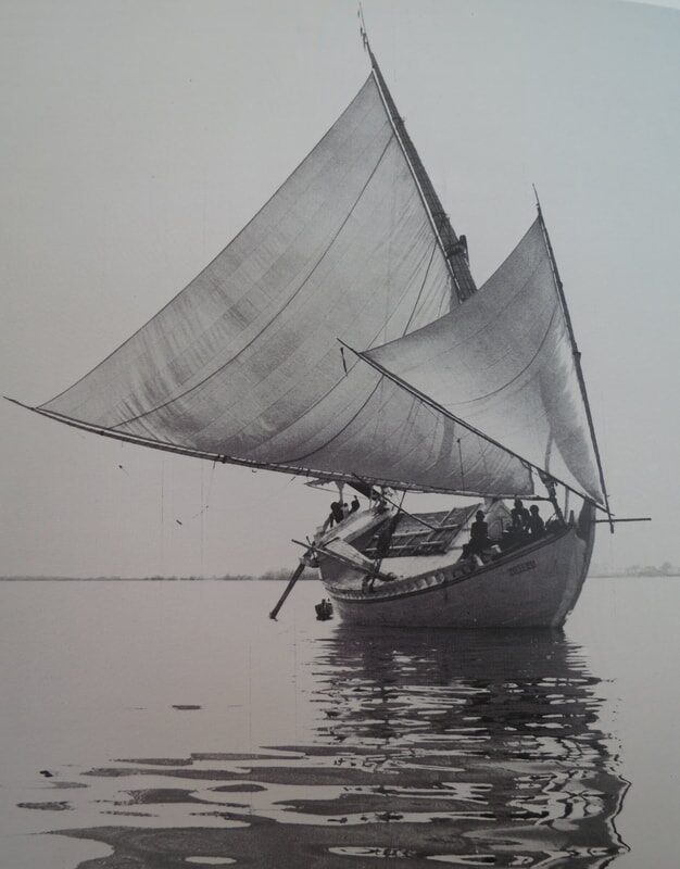 Black-and-white archival photo of a traditional Indonesian sailing boat at anchor, sails raised, crew aboard