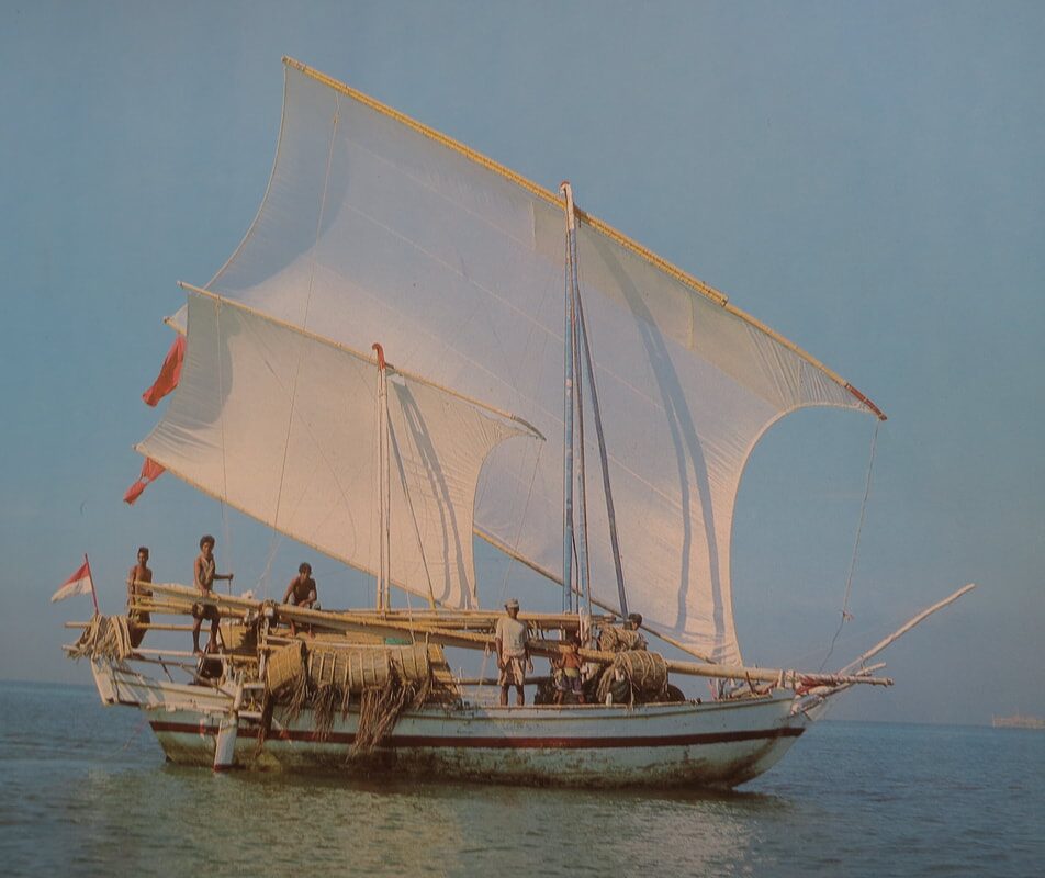 Color photo of a traditional Indonesian pinisi-style sailing ship under full white sail, crew working the deck