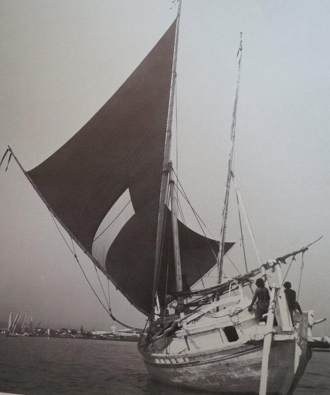Black-and-white archival photo of a two-masted Indonesian sailing vessel with a dark batwing sail, moored near a harbor skyline