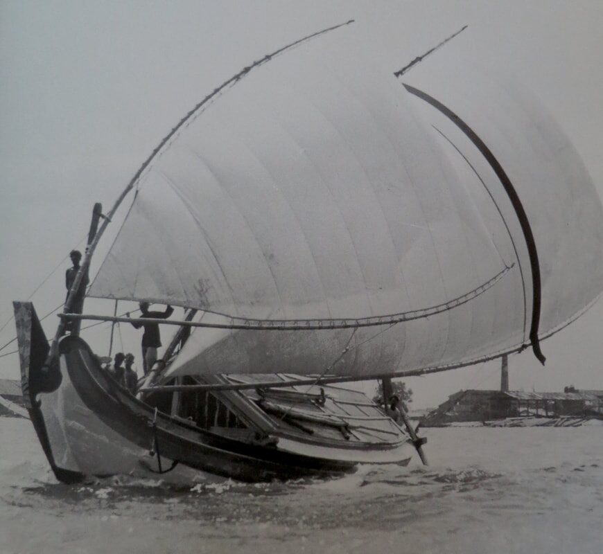 Archival photo of a traditional sailing boat heeling under a large curved sail near a shoreline of buildings