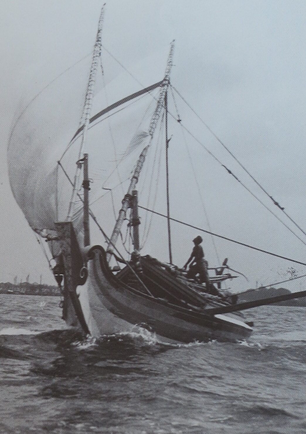 Archival photo of a tripod-masted Indonesian sailing vessel driving through the water under a full sail, one crewman standing at the bow