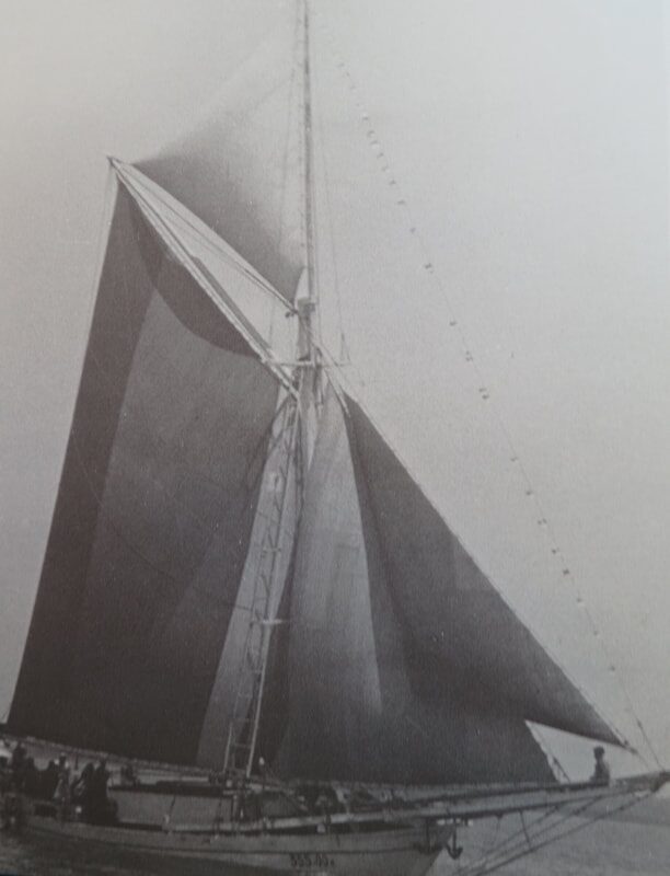 Archival photo of a traditional sailing boat's dark, tall sails silhouetted against the sky, crew at the rail