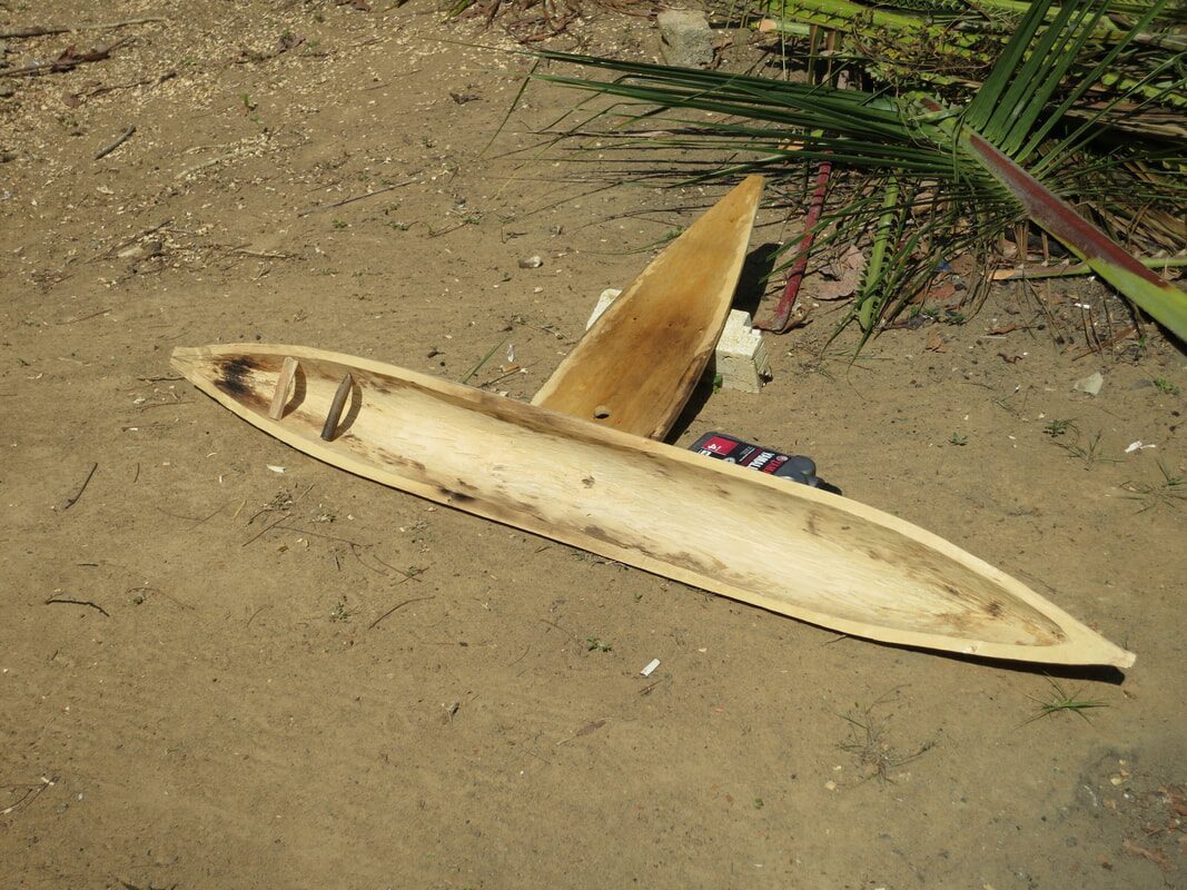 A freshly carved wooden jong hull and outrigger float resting on the ground beside palm fronds
