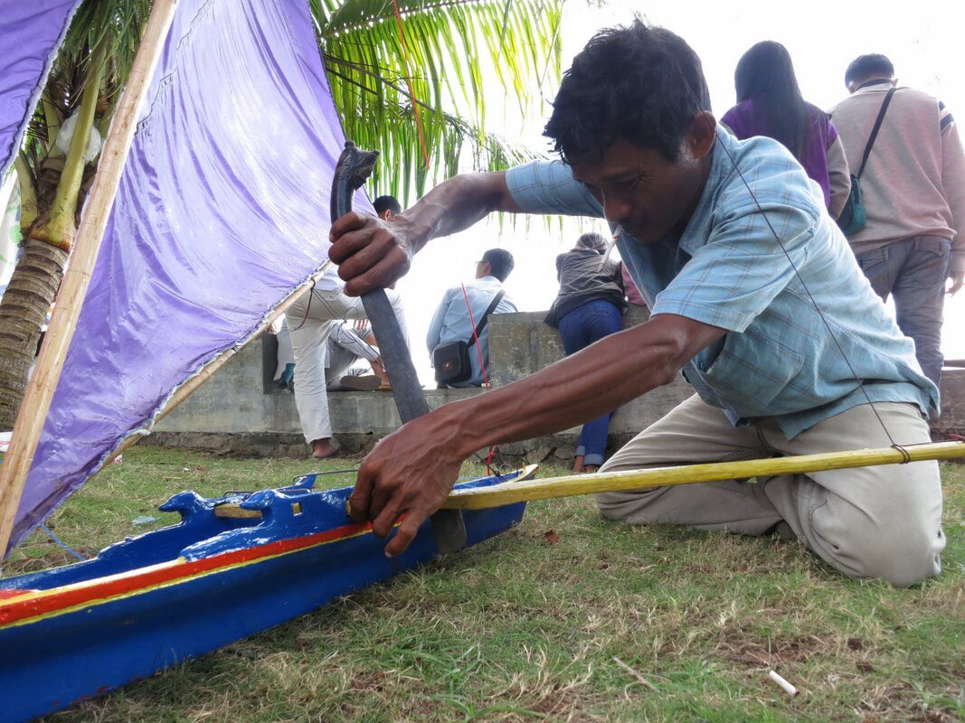 A man kneeling on grass, fitting rigging to a blue-hulled jong with a purple sail