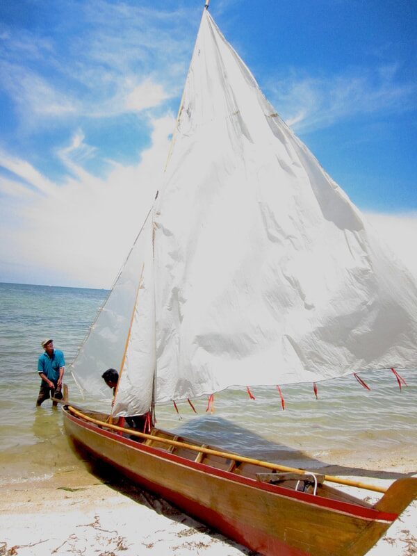 A traditional wooden sampan layar with a full white sail being launched from the beach, one man wading beside it and another aboard