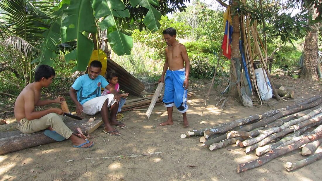 A group of men and a child carving and preparing wooden planks under trees, cut logs piled beside them