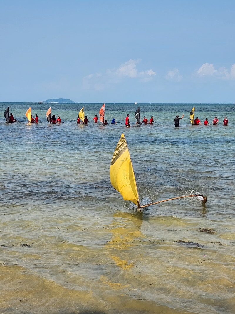 A yellow-sailed jong out front, with a line of young racers wading their own jongs through the shallows behind it