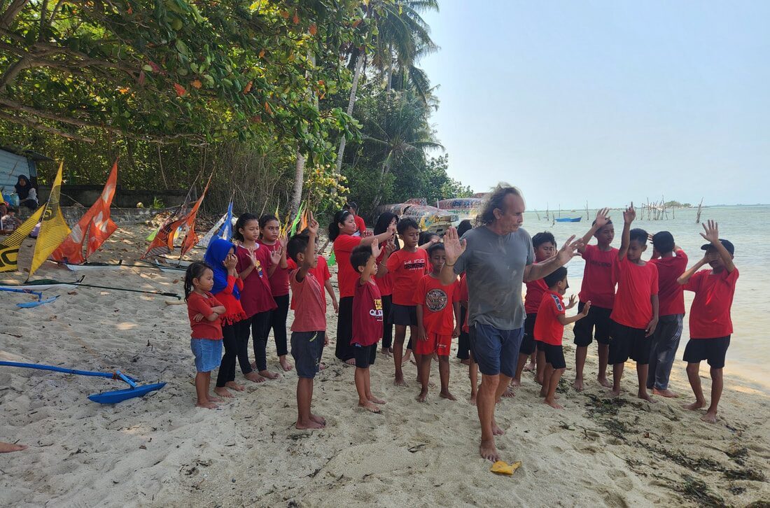 Michel leading kids in red Jong School shirts through a group warm-up on the beach, jong sails staked in the sand behind them