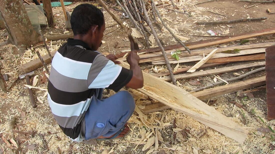 A man crouched carving a wooden plank with a hand tool, offcuts and sticks scattered around him