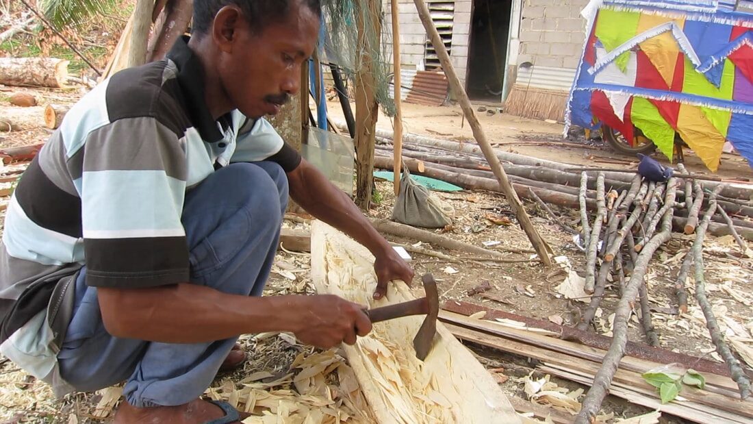 A man hammering a chisel into a wooden hull plank outdoors, colorful sail flags and stacked logs behind him