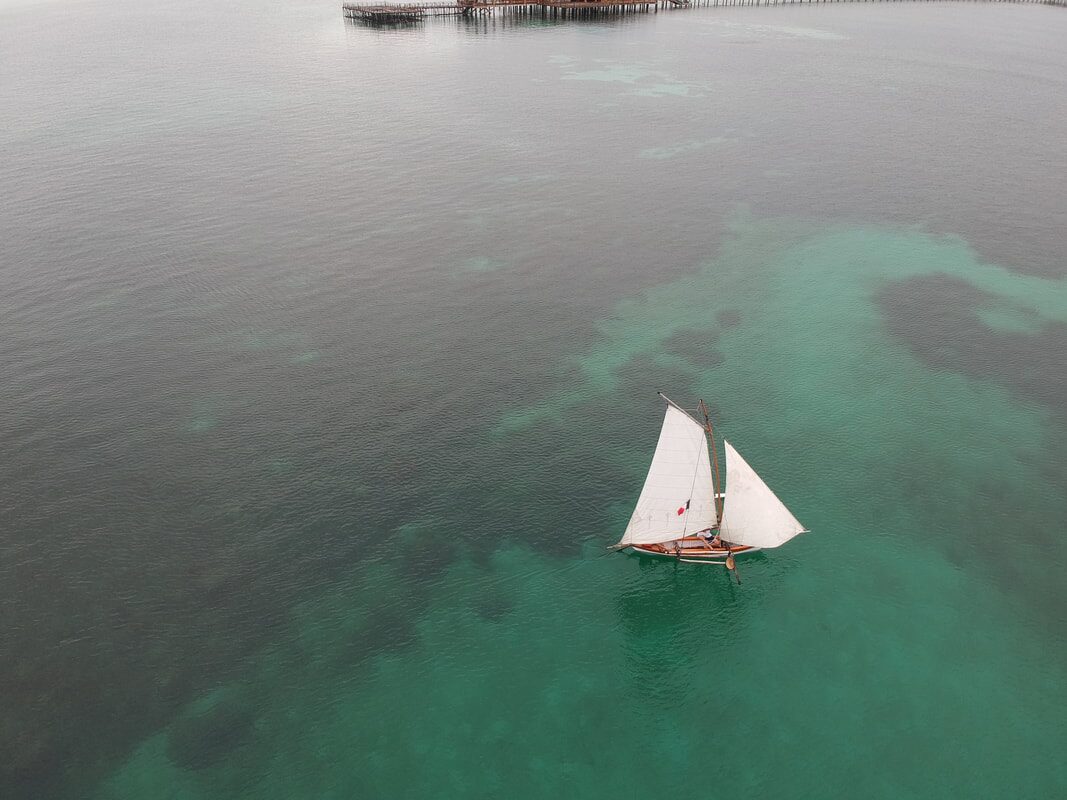 Aerial view of a white-sailed sampan layar sailing over a patchwork of turquoise and grey water, a stilt structure in the distance