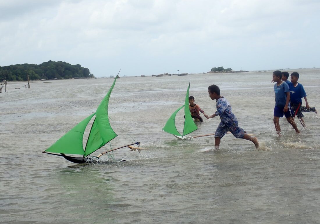 Boys racing green-sailed jongs through shallow water on the tidal flats, other kids running alongside