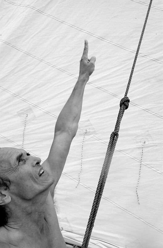 Black-and-white close-up of a shirtless man pointing up at the mast, sail cloth and rigging knots visible behind him