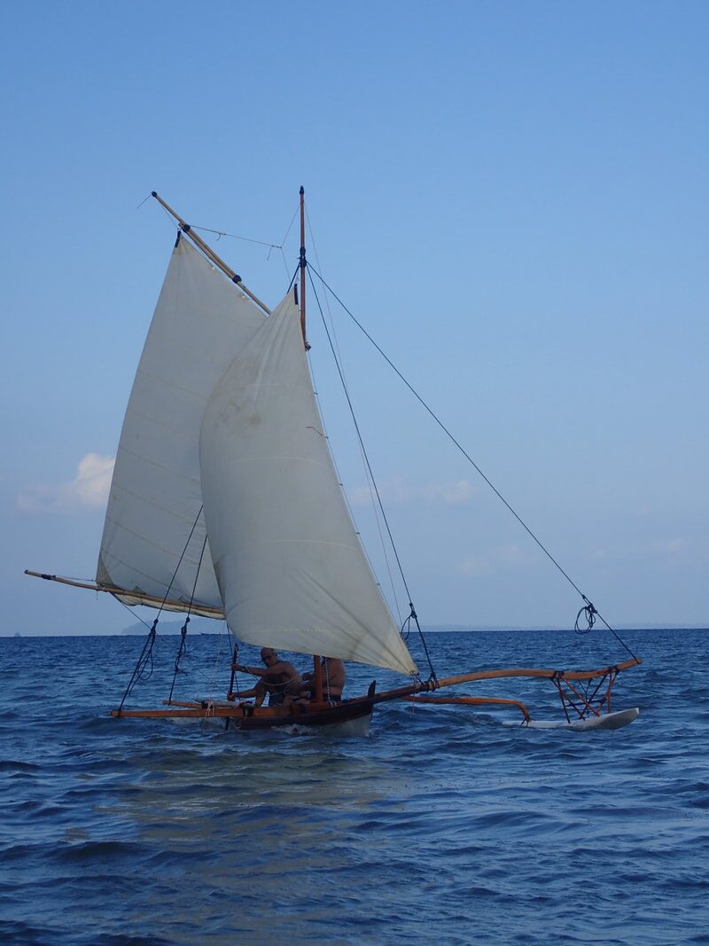 A wooden sampan layar under two white sails, crew steering, with an outrigger float trailing at its side